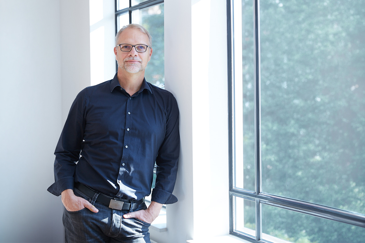 Andreas Ollmann lehnt am Fenster eines Lofts und schaut selbstbewusst in die Kamera