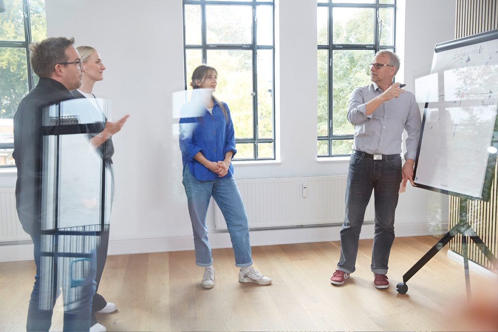 Ein Mann erklärt etwas am Flipchart, drei Kolleg:innen hören zu, in einem hellen Raum mit hohen Sprossenfenstern